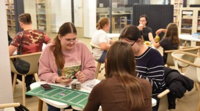 Na pierwszym planie trzy młode kobiety siedzą przy niewielkim stole, grając w grę planszową. Na stole leżą karty, żetony oraz zielona mata do gry. W tle widać inne osoby przy stolikach. Wnętrze jest jasne i nowoczesne, z wygodnymi krzesłami oraz regałami pełnymi rożnych gier planszowych.