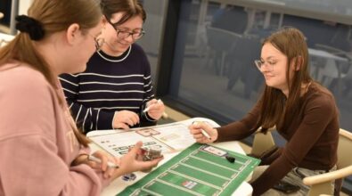 Trzy dziewczyny grają w grę planszową. Na stole leżą karty, żetony oraz zielona mata do gry.
