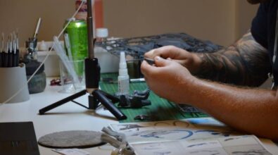 Zbliżenie na ręce osoby zajmującej się modelarstwem. Na stole znajdują się narzędzia, farby oraz elementy modeli, a obok stoi lampa oświetlająca miejsce pracy. Na biurku widoczny jest także laptop oraz różne akcesoria modelarskie, w tym pędzle i kleje.