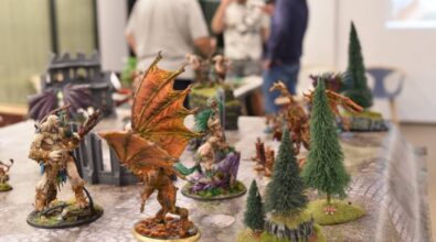 Na zdjęciu widać stół do gry bitewnej z bogato pomalowanymi figurkami fantasy. Na pierwszym planie znajduje się imponująca miniatura skrzydlatego demona o pomarańczowo-brązowych barwach oraz potężnego wojownika z maczugą. Wokół nich rozstawione są inne figurki przedstawiające fantastyczne postacie oraz elementy scenerii, takie jak drzewa, ruiny i wzgórza. W tle, lekko rozmyte, widoczne są osoby zaangażowane w rozgrywkę, rozmawiające i analizujące sytuację na planszy. Całość przypomina dynamiczną bitwę w świecie fantasy, prawdopodobnie w grze pokroju Warhammera lub innej strategicznej gry figurkowej.