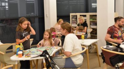 Na zdjęciu widać grupę osób grających w gry planszowe w przytulnej przestrzeni. Na pierwszym planie trzy osoby – dwie dziewczynki i młoda kobieta – grają w Monopoly. Jedna z dziewczynek stoi, trzyma kartę i wydaje się czytać jej treść, podczas gdy dwie pozostałe uczestniczki analizują sytuację na planszy. Na stole leżą pieniądze z gry, karty oraz różne napoje. Obok stoi kula ortopedyczna. W tle inne osoby również grają w gry planszowe, a na półkach za nimi widać różnorodne pudełka z planszówkami. Atmosfera jest swobodna i sprzyja wspólnemu spędzaniu czasu.