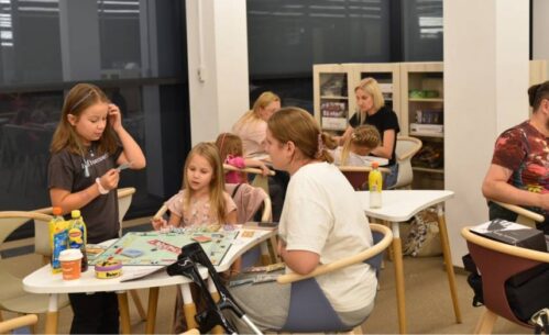 Na zdjęciu widać grupę osób grających w gry planszowe w przytulnej przestrzeni. Na pierwszym planie trzy osoby – dwie dziewczynki i młoda kobieta – grają w Monopoly. Jedna z dziewczynek stoi, trzyma kartę i wydaje się czytać jej treść, podczas gdy dwie pozostałe uczestniczki analizują sytuację na planszy. Na stole leżą pieniądze z gry, karty oraz różne napoje. Obok stoi kula ortopedyczna. W tle inne osoby również grają w gry planszowe, a na półkach za nimi widać różnorodne pudełka z planszówkami. Atmosfera jest swobodna i sprzyja wspólnemu spędzaniu czasu.