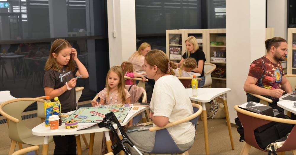 Na zdjęciu widać grupę osób grających w gry planszowe w przytulnej przestrzeni. Na pierwszym planie trzy osoby – dwie dziewczynki i młoda kobieta – grają w Monopoly. Jedna z dziewczynek stoi, trzyma kartę i wydaje się czytać jej treść, podczas gdy dwie pozostałe uczestniczki analizują sytuację na planszy. Na stole leżą pieniądze z gry, karty oraz różne napoje. Obok stoi kula ortopedyczna. W tle inne osoby również grają w gry planszowe, a na półkach za nimi widać różnorodne pudełka z planszówkami. Atmosfera jest swobodna i sprzyja wspólnemu spędzaniu czasu.