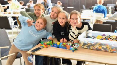 Grupa młodych uczennic pozuje do zdjęcia przy stole, na którym stoi duże pudełko z klockami LEGO. Obok pudełka leży gotowa budowla. W tle widać inne dzieci siedzące przy stolikach.