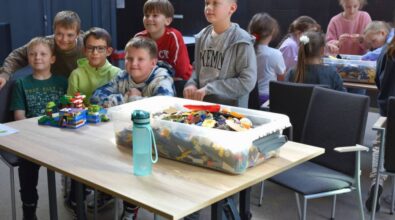 Uczniowie pozują do zdjęcia przy stoliku, na którym leży zbudowana konstrukcja. Na stoliku widać również duże pudełko z klockami, a w tle inną grupę dzieci.