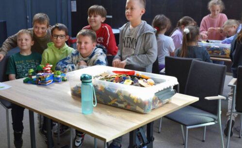 Uczniowie pozują do zdjęcia przy stoliku, na którym leży zbudowana konstrukcja. Na stoliku widać również duże pudełko z klockami, a w tle inną grupę dzieci.