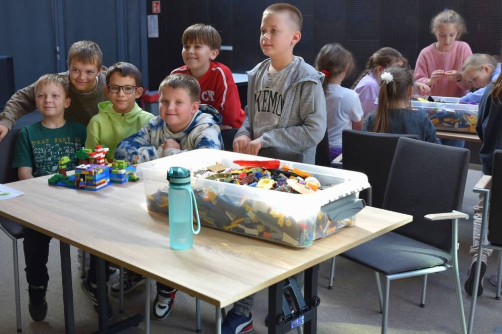 Uczniowie pozują do zdjęcia przy stoliku, na którym leży zbudowana konstrukcja. Na stoliku widać również duże pudełko z klockami, a w tle inną grupę dzieci.