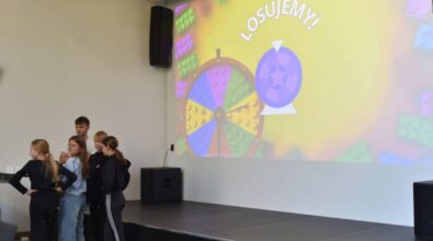 Prowadzący wraz z grupką uczestników stoi przy mównicy obok ekranu projekcyjnego. Na ekranie widać slajd z kolorowym kołem zbudowanym z klocków LEGO i napis 