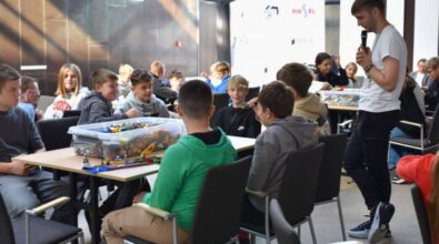 Uczniowie siedzą przy stolikach, na których stoją pudełka z klockami. Osoba prowadząca przemawia do mikrofonu.