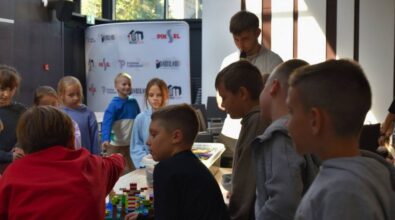 Uczniowie siedzą przy stolikach, na których stoją pudełka z klockami. Osoba prowadząca przemawia do mikrofonu.