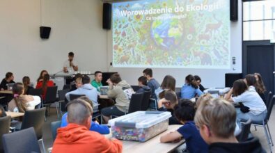 Uczniowie siedzą przy stolikach, na których stoją pudełka z klockami. Osoba prowadząca przemawia do mikrofonu, w tle widać ekran projekcyjny i slajd z napisem 