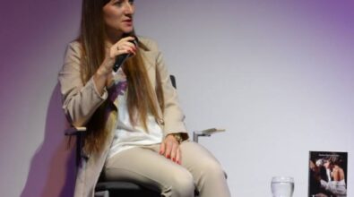 Na zdjęciu widzimy autorkę Paulinę Marię Kowalską siedzącą na krześle na tle fioletowego oświetlenia. Jest elegancko ubrana w jasny garnitur, a w dłoni trzyma mikrofon. Na małym stoliku obok niej znajduje się promowana książka oraz szklanka z wodą.