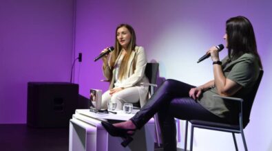 Na zdjęciu widzimy dwie kobiety siedzące na scenie podczas rozmowy. Obie trzymają mikrofony. Kobieta po lewej stronie ubrana jest w elegancki, jasny garnitur, a po prawej stronie druga osoba ma na sobie bardziej casualowy strój z zieloną koszulą i czarnymi skórzanymi spodniami. Między nimi na stoliku znajduje się książka.