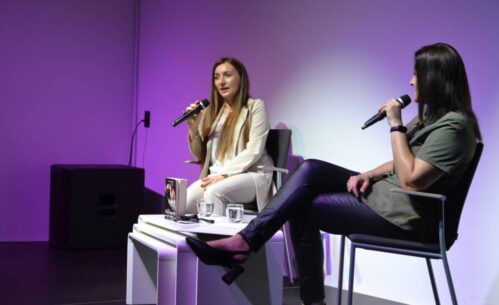 Na zdjęciu widzimy dwie kobiety siedzące na scenie podczas rozmowy. Obie trzymają mikrofony. Kobieta po lewej stronie ubrana jest w elegancki, jasny garnitur, a po prawej stronie druga osoba ma na sobie bardziej casualowy strój z zieloną koszulą i czarnymi skórzanymi spodniami. Między nimi na stoliku znajduje się książka.
