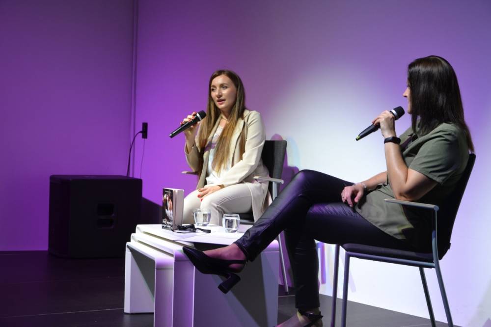 Na zdjęciu widzimy dwie kobiety siedzące na scenie podczas rozmowy. Obie trzymają mikrofony. Kobieta po lewej stronie ubrana jest w elegancki, jasny garnitur, a po prawej stronie druga osoba ma na sobie bardziej casualowy strój z zieloną koszulą i czarnymi skórzanymi spodniami. Między nimi na stoliku znajduje się książka.