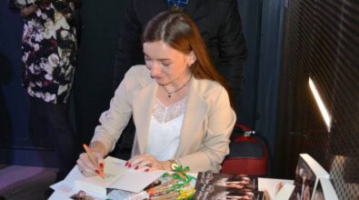 Autorka siedzi przy stoliku i podpisuje egzemplarze swojej książki. Obok niej stoją ludzie, czekając na swoją kolej. Na stoliku leży kilka egzemplarzy książki pod tytułem 'Trzy minuty do północy', a także długopisy i zakładki.