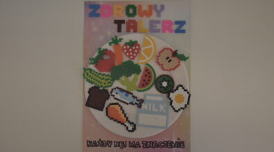 Praca konkursowa w formie plakatu przedstawiająca umieszczony centralnie biały papierowy talerz z różnymi produktami, warzywa, owoce, ryba, kurczak, chleb, jajko. Elementy z sprasowanych sklejonych koralików. Na dole czarny napis „KAŻDY KĘS MA ZNACZENIE”. Na górze kolorowy napis „ZDROWY TALERZ”. Tło różowe z rozbłyskami.