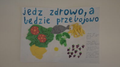 Praca konkursowa w formie plakatu przedstawiająca umieszczone centralnie różne produkty, sałata z bibuły, pomidory, żółty produkt, jajko z kartek, namalowany brokuł i jeżyny, ryba z filcu i makaron z pianki. Z lewej strony małe napisy dotyczące zdrowego żywienia. Na górze niebieski napis „jedz zdrowo, a będzie przebojowo”. Tło białe.