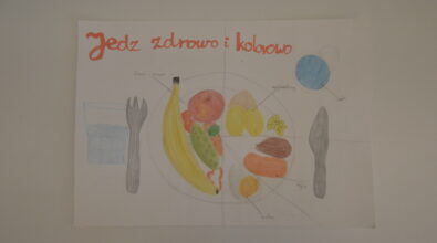 Praca konkursowa w formie plakatu przedstawiająca umieszczony centralnie podzielony na 3 części biały talerz z różnymi produktami, warzywa, owoce, produkty zbożowe, mięso, jajko. Produkty z małymi podpisami. Po bokach sztućce i napoje w szklance i kubku. Na górze pomarańczowy napis „Jedz zdrowo i kolorowo”. Praca wykonana kredkami. Tło białe.