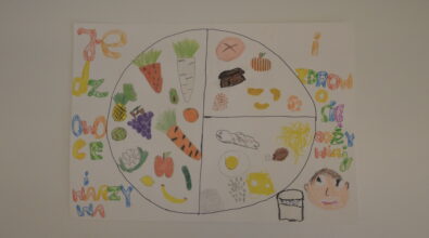 Praca konkursowa w formie plakatu przedstawiająca umieszczony centralnie podzielony na 3 części biały talerz z różnymi produktami, warzywa, owoce, produkty zbożowe, nabiał, jajko, mięso. Po lewej stronie kolorowy napis „Jedz OWOCE I WARZYWA”. Po prawej stronie kolorowy napis „i ZDROWO SIĘ ODŻYWIAJ”. W prawym dolnym rogu twarz dziecka i karton mleka. Praca wykonana kredkami. Tło białe.