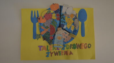 Praca konkursowa w formie plakatu przedstawiająca umieszczony centralnie pomalowany na niebiesko talerz podzielony na 3 części z różnymi produktami, warzywa, nabiał, ryba, mleko, produkty zbożowe, prawdziwa kasza, makaron, reszta wykonana z naklejonych pokolorowanych kartek. Po bokach niebieskie naklejone sztućce. Pod talerzem kolorowy napis „TALERZ ZDROWEGO ŻYWIENIA” z literami narysowanymi i wyklejonymi kulkami z bibuły. Tło żółte.