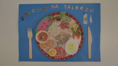 Praca konkursowa w formie plakatu przedstawiająca różowy talerz z różnymi produktami, warzywa, owoce, jajko, ryba z wklejonych kartek, prawdziwe orzechy, płatki owsiane, pomarańcza. Po bokach drewniane sztućce. Na górze kolorowy napis „ZDROWIE NA TALERZU”. Tło niebieskie.