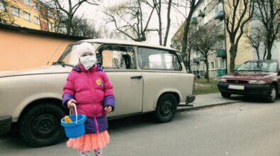 Mała dziewczynka ubrana w grubą różową kurtkę i pomarańczową spódniczkę trzyma w ręce niebieskie wiaderko, a na twarzy ma maseczkę ochronną. Stoi przodem do obiektywu. Tuż za nią stoi stary samochód w jasnym kolorze, a dalej drugi – bordowy. W tle dwa bloki mieszkalne.