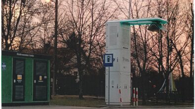 Wysoka, biała kolumna zakończona u góry wysięgnikiem z panelem fotowoltaicznym lub elementem oświetleniowym. Obok stoi niebieski znak informacyjny. Po lewej stronie znajduje się zielony kontener techniczny z czarnymi drzwiami i naklejkami ostrzegawczymi. Całość znajduje się przy asfaltowej drodze. W tle widoczne są drzewa bez liści oraz niebo w odcieniach różu i fioletu.
