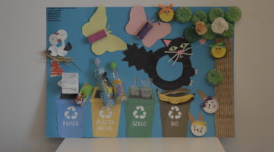 Kolorowa praca plastyczna w formie plakatu wykonana z materiałów z recyklingu. Na plakacie cztery pojemniki do segregowania śmieci wykonane z papieru. Nad pojemnikami przyklejone papierowe opakowania, plastikowe butelki, wazony z folii bąbelkowej i skórka od banana. Po prawej stronie drzewo z papieru i plastikowych butelek pomalowanych na zielono, ptaszki i kotki wykonane z metalowych nakrętek i papieru oraz czarny kot z papieru. Po lewej stronie gniazdo ze słomy z bocianami z płatków kosmetycznych oraz motyle z papieru.