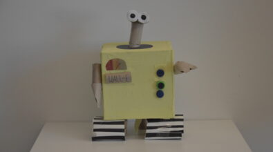 Praca plastyczna przedstawiająca robota. Tułów wykonany z sześciennego kartonu obklejonego żółtym papierem. Głowa z rolek po papierze z dużymi oczami z białego i czarnego papieru. Ręce z rolek po papierze. Gąsienice z rolek po papierze i pasków białego i czarnego papieru. Na tułowiu po prawej stronie przyciski z trzech plastikowych nakrętek, po lewej stronie wskaźnik mocy z papieru i napis WALL-E.