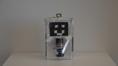 Praca plastyczna przedstawiająca robota wykonanego z materiałów recyklingowych. Całość wykonana z otwieranego pudełka owiniętego folią aluminiową. Na przodzie znajduje się kwadratowa twarz z czarnego papieru, z naklejonymi oczami i ustami z białego papieru. Ręce wykonane z patyczków owiniętych folią aluminiową trzymają puszkę Pepsi ze słomką.