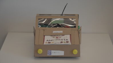 Praca plastyczna przedstawiająca samochód wykonany z materiałów recyklingowych. Korpus posklejany z elementów wyciętych z kartonu. Przednia szyba z przezroczystej folii z czarnymi wycieraczkami. Na dachu antena z czarnego plastiku. Koła wykonane z białych plastikowych nakrętek. Na przedniej masce biały kartonik z napisem. Pod uniesionym kartonikiem widoczna komora silnika z elementami wykonanymi z tektury, papieru, słomek i drobnych plastikowych części. Wlew do baku z zielonej plastikowej nakrętki. Drzwi samochodu są otwierane. Tablica rejestracyjna wykonana z białej kartki papieru. Przednie światła z żółtych plastikowych nakrętek, tylne z czerwonych i pomarańczowych plastikowych nakrętek. Wewnątrz samochodu widoczna kierownica z dużej czerwonej plastikowej nakrętki oraz zielone siedzenia.