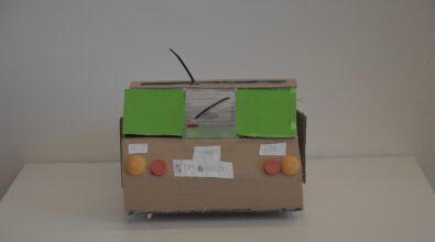 Praca plastyczna przedstawiająca samochód wykonany z materiałów recyklingowych. Przedstawiony tył auta. Korpus posklejany z elementów wyciętych z kartonu. Tylna szyba z przezroczystego plastiku z czarną wycieraczką. Tablica rejestracyjna wykonana z białej kartki papieru. Tylne światła z żółtych plastikowych nakrętek, tylne z czerwonych i pomarańczowych plastikowych nakrętek.
