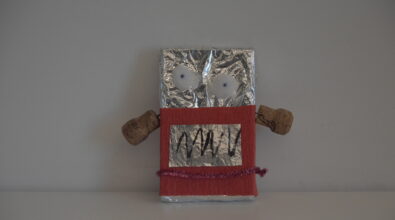 Praca plastyczna przedstawiająca stworka z sześciennego kartonika, obklejonego folia aluminiową. Duże oczy wykonane z waty i szpilek z niebieską główką. Ręce z korków od butelek. Dolna część tułowia ozdobiona czerwoną krepiną, folią aluminiową i różowym drucikiem kreatywnym.