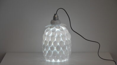 Lampa w owalnym kształcie wykonana z białych plastikowych łyżeczek. Podłączona do prądu kablem. Dająca światło.