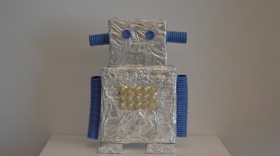 Praca plastyczna przedstawiająca robota wykonanego z materiałów recyklingowych. Tułów i głowa z dwóch sześciennych kartonów owiniętych folią aluminiową. Głowa mniejsza od tułowia. Oczy z niebieskich plastikowych nakrętek. Uszy i ręce z niebieskich rurek karbowanych, nogi z małych, płaskich kartoników owiniętych folia aluminiową. Na środku tułowia złoty, plastikowy element z piętnastoma uwypukleniami.