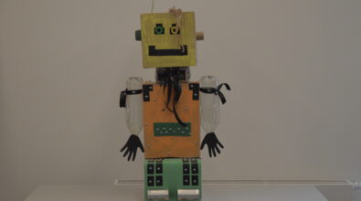 Robot wykonany z materiałów z recyklingu. Nogi, tułów, szyja i głowa wykonane z kartonów pomalowanych na kolor zielony, pomarańczowy i żółty. Usta z czarnego papieru, oczy z czarnego papieru i nakrętek plastikowych, jedno ucho z dużej, zielonej, plastikowej nakrętki, drugie ucho z rolki po papierze. Na głowie kosmyk włosów z papieru i antenka z dwiema fioletowymi kuleczkami. Ręce z plastikowych butelek, dłonie z czarnego papieru. Stopy z puszek po konserwach. Na szyi czarna kokarda. Tułów i nogi obklejone czarnymi prostokątami i naklejkami ruchomych oczu.