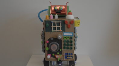 Praca plastyczna przedstawiająca robota wykonanego z materiałów recyklingowych, przypominającego dużą kostkę sensoryczno-edukacyjną. Robot ma korpus z kartonowego pudełka ustawionego na kołach wykonanych z plastikowych butelek i papierowych kubeczków. Z przodu znajduje się wiele elementów manipulacyjnych i dekoracyjnych: klawiatura z przyciskami z plastikowych nakrętek, okrągłe pokrętło z plastikowych kubeczków, małe okienka z patyczków, kolorowe guziki z nakrętek oraz różnorodne elementy z plastiku i tektury. Na górze umieszczono głowę robota z dużymi oczami z kulek oraz antenkami wykonanymi ze słomek.